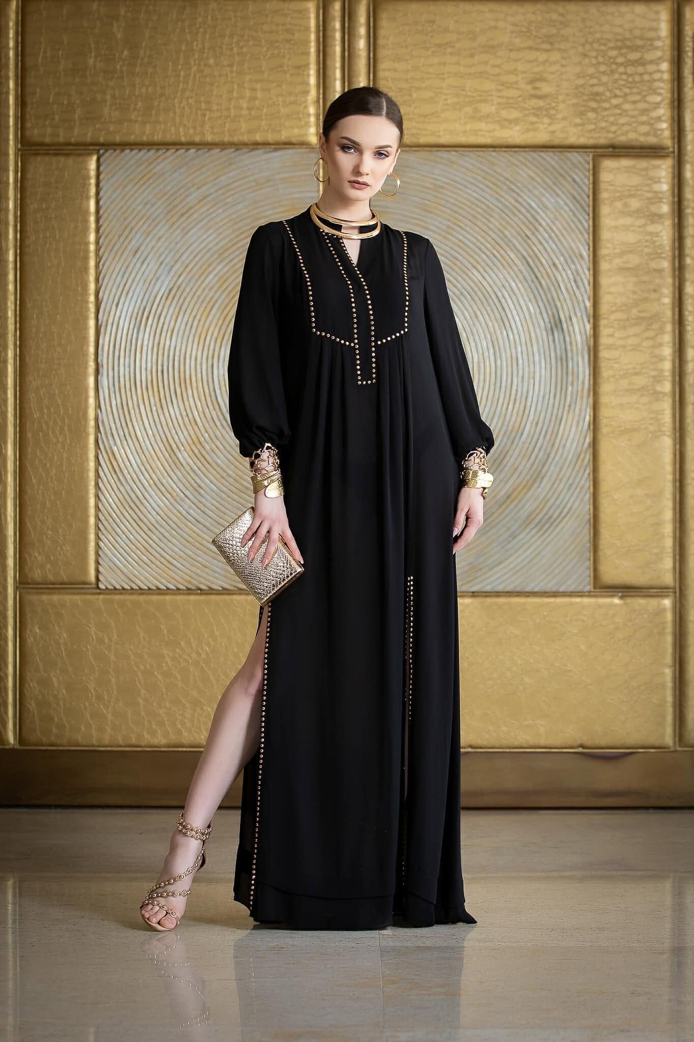 Model purtând rochia SELENA, rochie neagră tip caftan cu studuri aurii, în fața unui perete auriu.